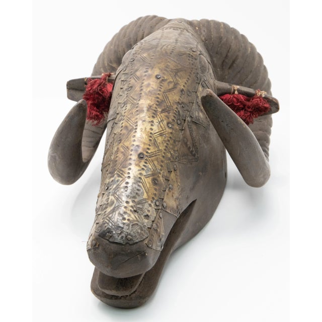 Carved wooden ram mask with brass overlay. Horns are carved wrapping the head on either side. Ears have tassels in them...