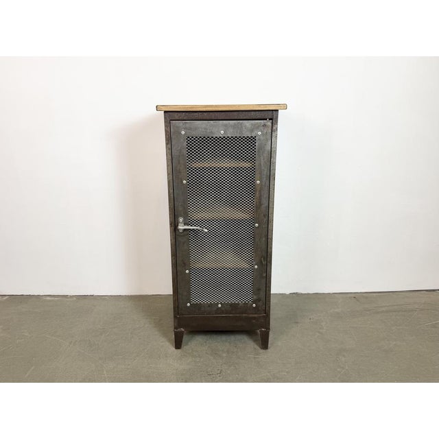 An iron cabinet from the 1960s features an iron structure, mesh doors, a wooden top and three wooden shelves inside the...