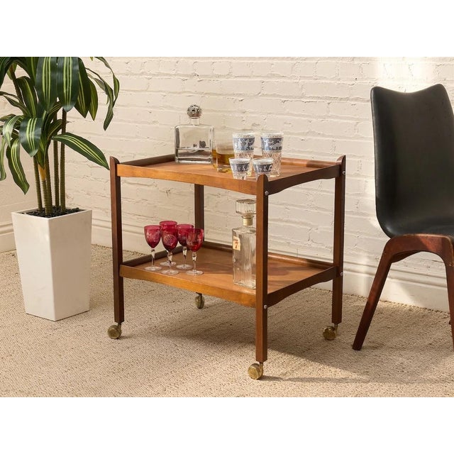 A handsome vintage two-tier bar cart featuring a warm wood finish and a clean, understated mid-century silhouette. Two...