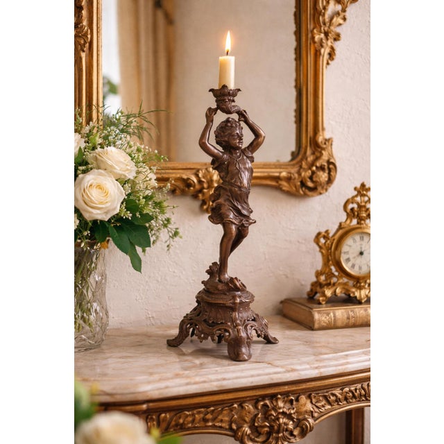 A striking antique-style figural candle holder featuring a joyful cherub gracefully posed atop an ornate Rococo-inspired...