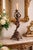 A striking antique-style figural candle holder featuring a joyful cherub gracefully posed atop an ornate Rococo-inspired...