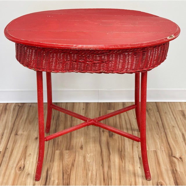 Vintage wicker table with oval slatted wood top in a weathered bold red paint. Four wrapped legs attached by x-base...