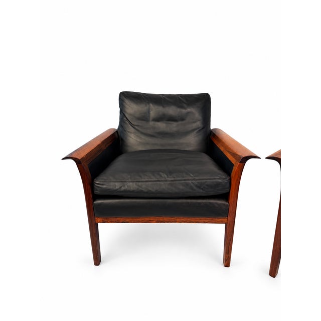 A pair of Hans Olson rosewood and black leather lounge chairs, unmarked. Shaped wooden arms and exposed frames trace the...