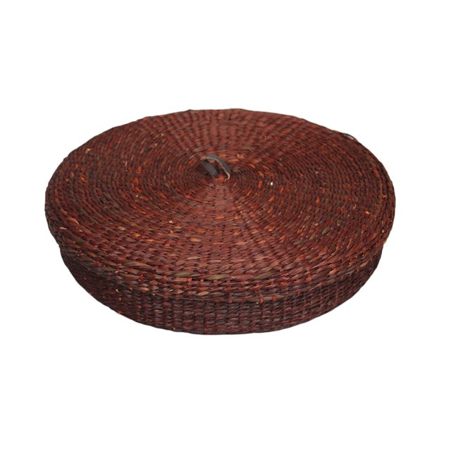 A set of six circular woven raffia placemats with matching woven storage basket. Mats are a deep reddish-brown color with...