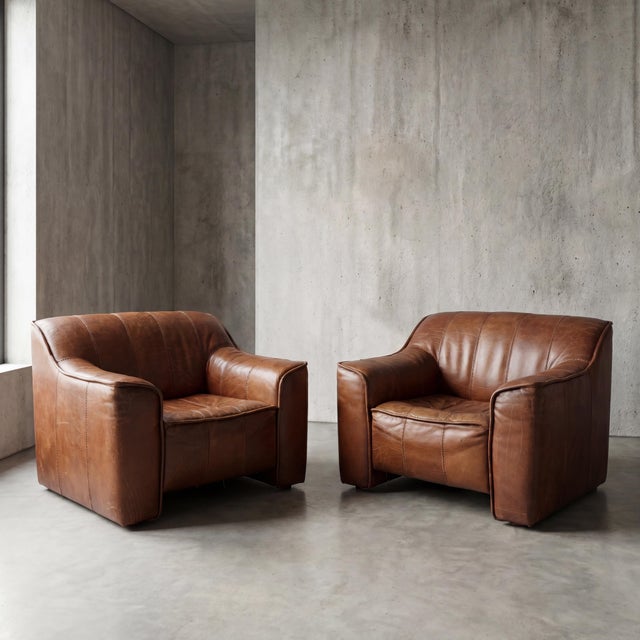 Thick, deeply patinated cognac hide drapes across the low-slung frames of these commanding club chairs, offering an...