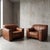 Thick, deeply patinated cognac hide drapes across the low-slung frames of these commanding club chairs, offering an...
