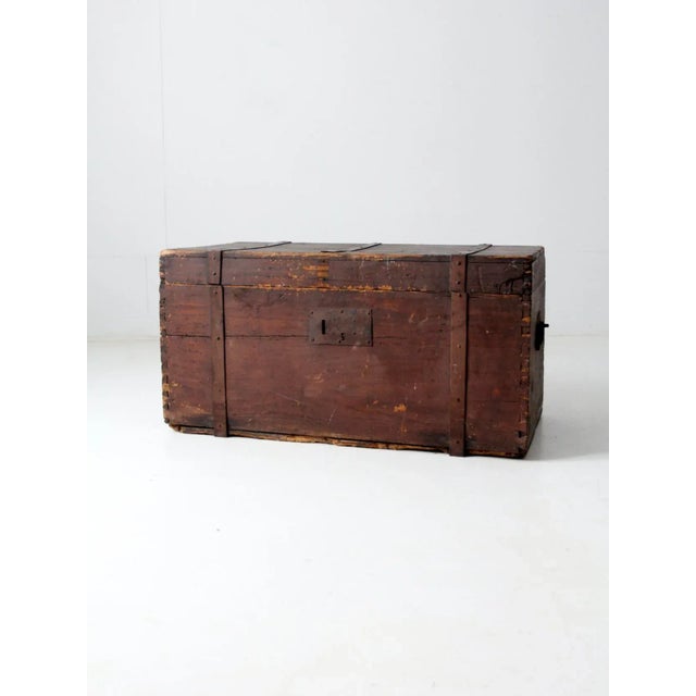 This is an antique wooden trunk. It features a flat top with metal bracing straps and metal handles on the sides. A heavy...