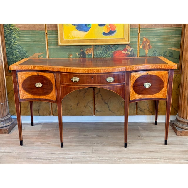This early 19th C. English Hepplewhite sideboard has a serpentine mahogany top with a wide satinwood crossbanded border...