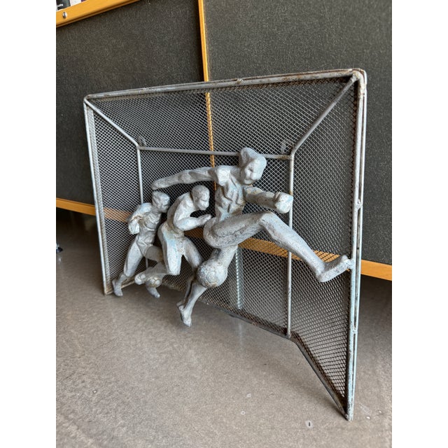 This vintage metal wall sculpture depicts three athletic figures in high relief against a wire mesh soccer goal frame....
