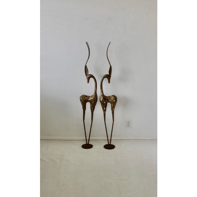 A pair of tall, stylized bronze-toned metal gazelle sculptures with slender legs and elegant curved horns. Individually...