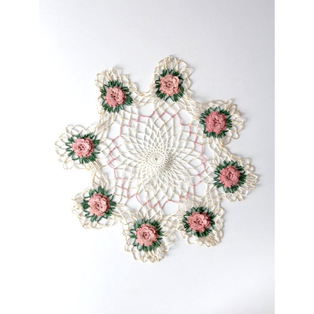 A vintage crocheted doily featuring a ring of raised pink floral motifs set against an open lace ground. Soft tones of...