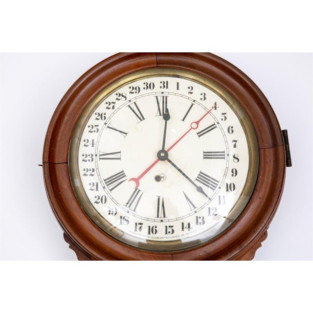 An antique wall clock in a mahogany case with a large round bezel, white dial with black hour and minute hands, bold black...
