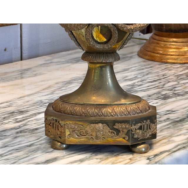 A brass mantel clock in the form of a two-handled urn, with the clock face set into the body of the vase. The handles...