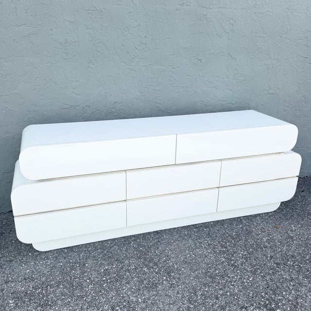 A striking postmodern dresser defined by its fluid waterfall silhouette and luminous white gloss laminate finish. Softly...