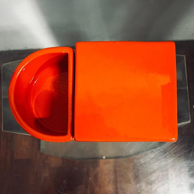 Small orange ceramic storage box, consisting of two containers, one with lid, the other crescent-shaped, open. Produced by...