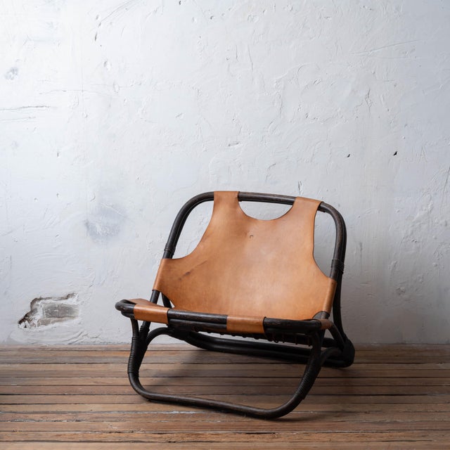 A sculptural mid-century lounge chair in rattan with a dark bent-frame structure and a tan leather sling seat and back....
