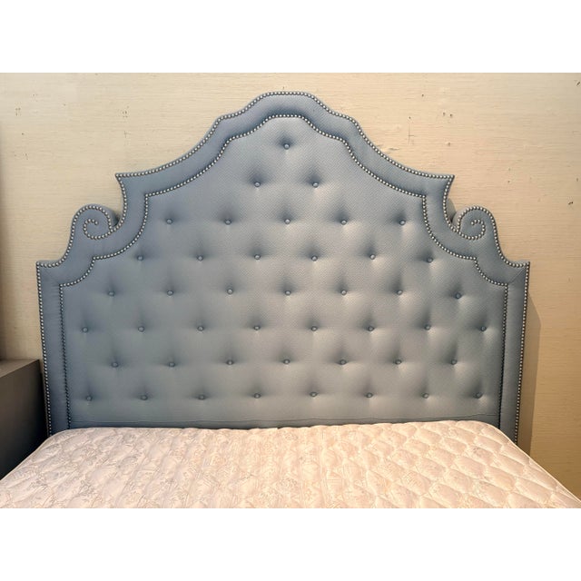 Glamorous bed features a dramatic, shapely headboard fully upholstered in a soft blue-grey fabric, finished with deep...