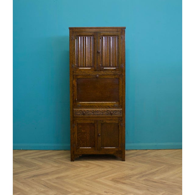 A 1920s oak kitchen larder cupboard in the Tudor Revival style, featuring vented upper doors, a central fall-front...
