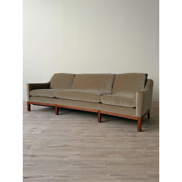 Mid-century sofa with a subtly curved profile, upholstered arms, and an exposed wood base. The frame supports three loose...