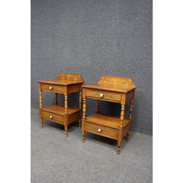 Pair of Hitchcock maple nightstands with two drawers, lower shelf, turned legs, and decorated backsplashes. Each table...