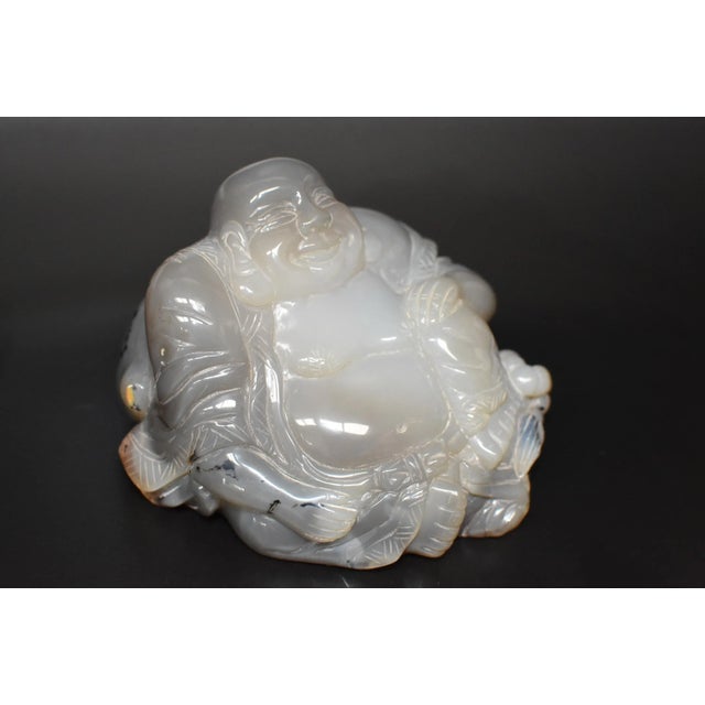 A hand-carved, solid agate Buddha. He is depicted sitting with his robe open, cinched with a bow under his belly, an...