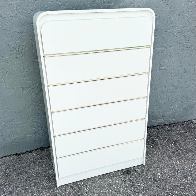 A sleek postmodern highboy dresser, rendered in a crisp white gloss laminate with a striking waterfall silhouette. The...