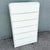 A sleek postmodern highboy dresser, rendered in a crisp white gloss laminate with a striking waterfall silhouette. The...