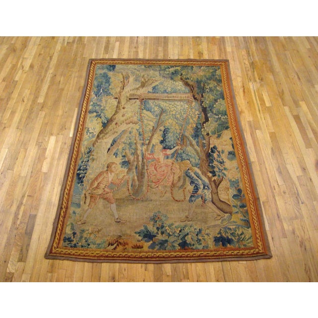 A Flemish rustic tapestry from the 18th century, depicting a charming scene with two young noblemen pulling a young damsel...
