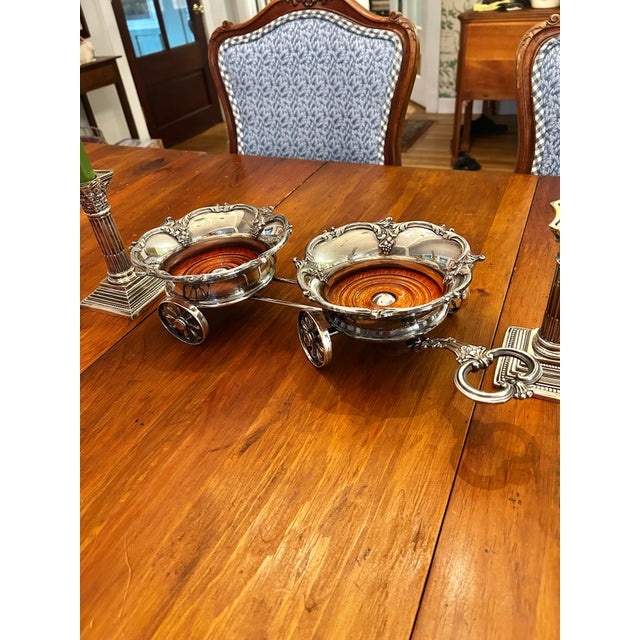 This wine bottle or decanter trolley features two elaborate silver-plated holders on functioning wheels with a long...