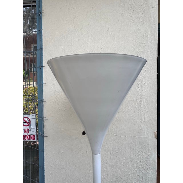 This 1970s torchiere is a sober sculptural floor lamp. A deep white trumpet shade rises from a slender tapered stem on a...