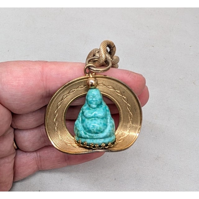 1950s goldtone etched circle with cut out center and bent bottom with a carved faux-jade buddha charm on a chain with...