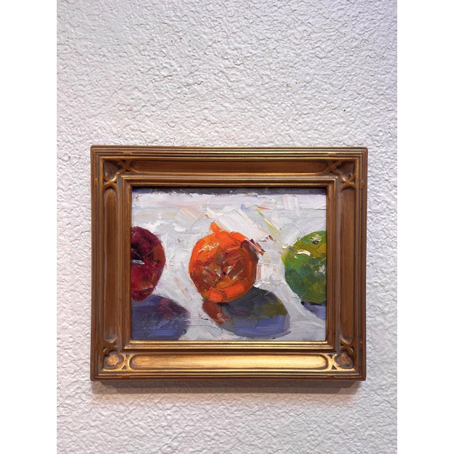This oil painting features three vibrant fruitsan orange, a red apple, and a green appleset against a softly textured...