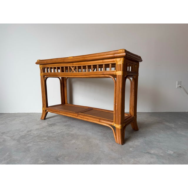 A stylish vintage coastal rattan console table featuring a two-tier design with woven cane panels and subtle leather-...