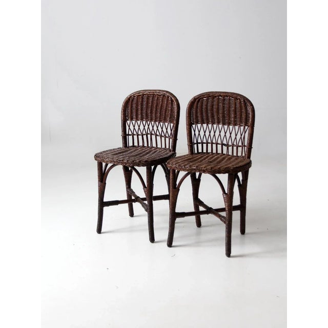 A pair of antique wicker chairs distinguished by their low backs, rounded seats, and distinctive thatched detailing. The...
