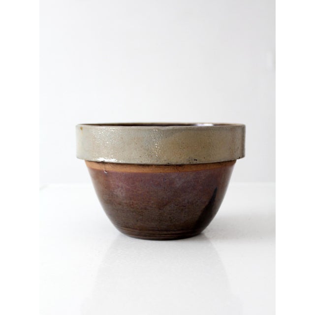 This antique American stoneware mixing bowl features a classic utilitarian form with a soft buff-gray rim and rich brown...