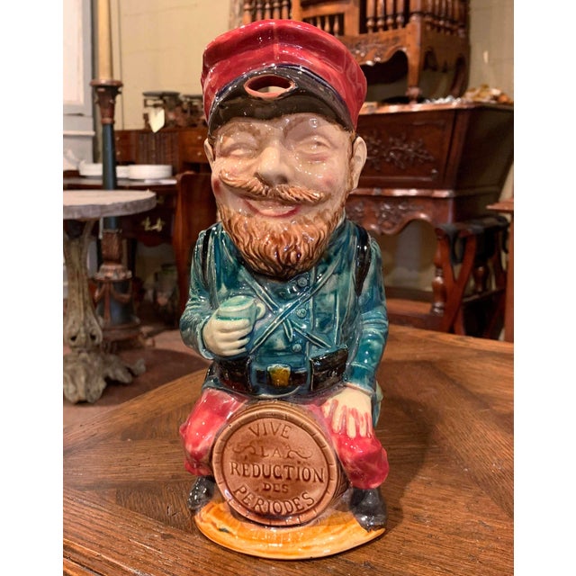 Colorful and highly expressive, this antique barbotine pitcher depicts a Belgian soldier seated confidently upon a barrel....