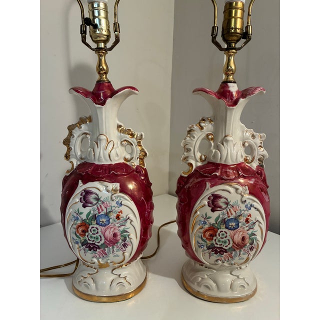 A Pair of vintage mid-century ceramic table lamps featuring ornate rococo scrollwork, iridescent red glaze, and hand-...
