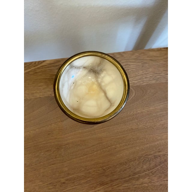 A small footed bowl turned from alabaster, banded in brass at the rim. It stands on its own short pedestal, so it reads...