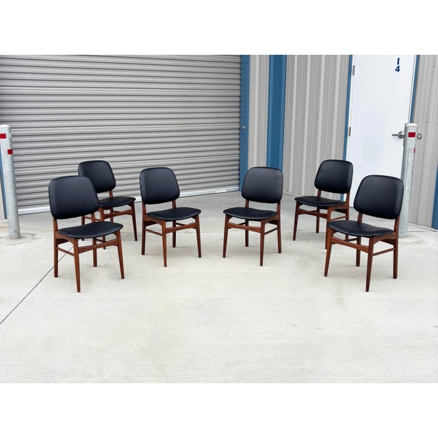 This exceptional set of six mid-century modern dining chairs beautifully pairs warm, teak frames with sleek black vinyl...