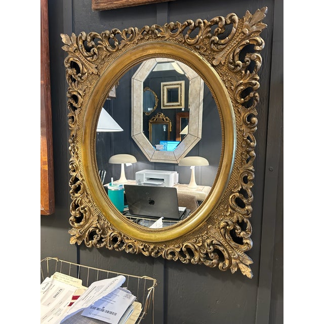 Vintage oval mirror with an ornate carved wood frame finished in gold leaf. Features intricate scrollwork and layered...