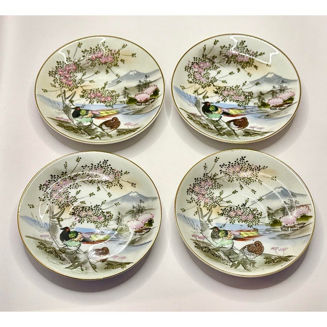 Four hand‑painted Japanese Kutani saucers, each featuring a scenic landscape with birds, blossoms, and soft mountain...