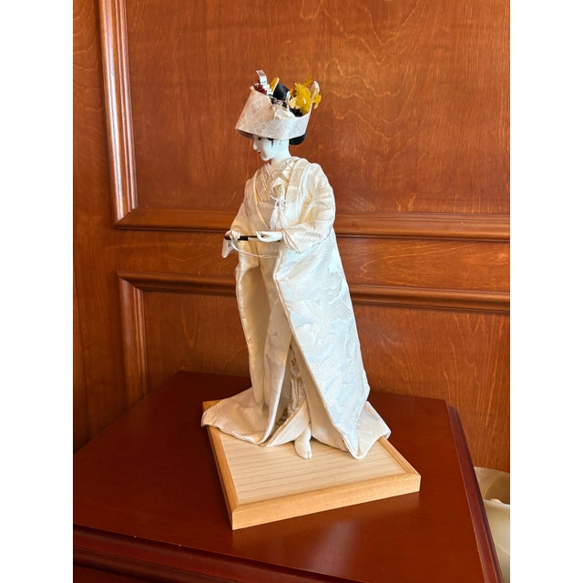 A graceful vintage Japanese ceremonial figure dressed in a flowing ivory-toned kimono, presented on a natural wood base....