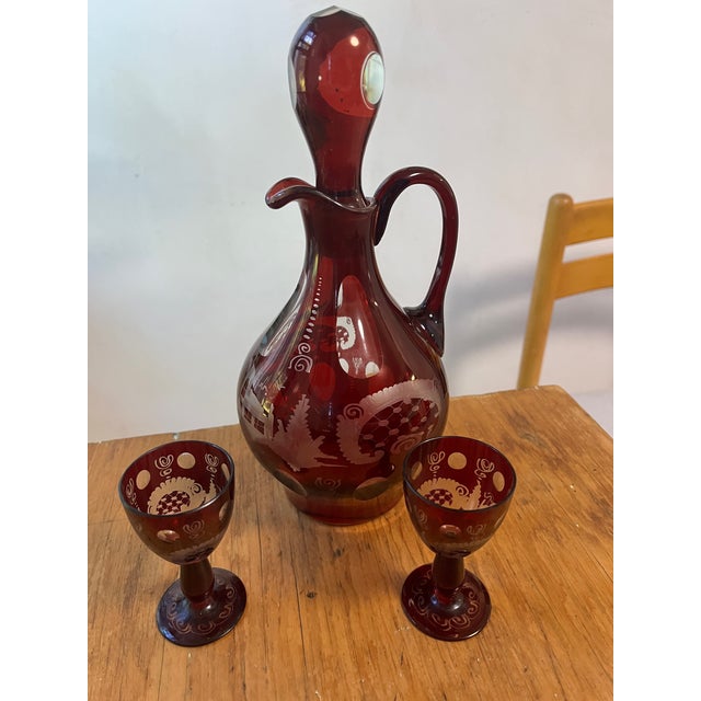 A magnificent antique bohemian etched glass decanter with two matching glasses. Cranberry red to clear glass bottle with...
