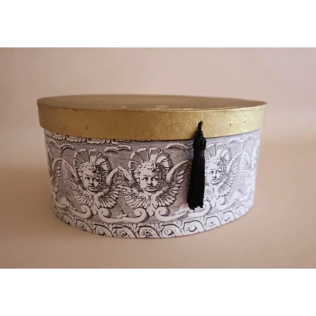 Oval gold lidded paper box decorated with black and white cherub print. Front of gold lid embellished with black tassel.