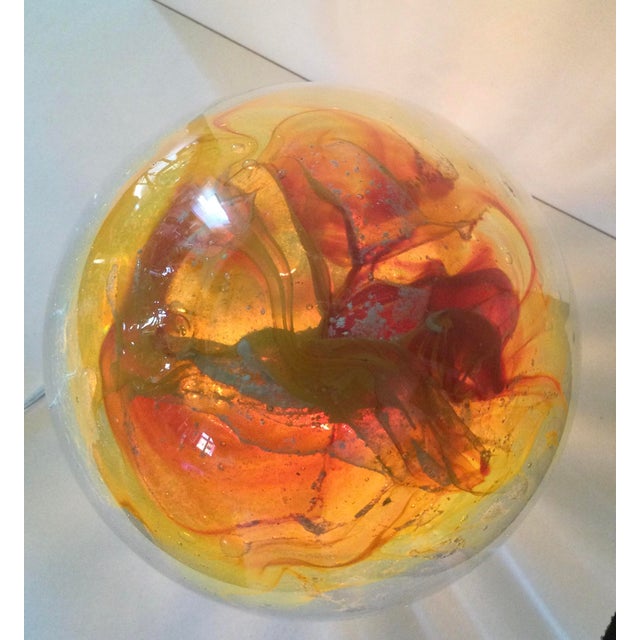 The Egg of the Sun table lamp is crafted from blown glass, blending red-orange hues with luminescent minerals that charge...