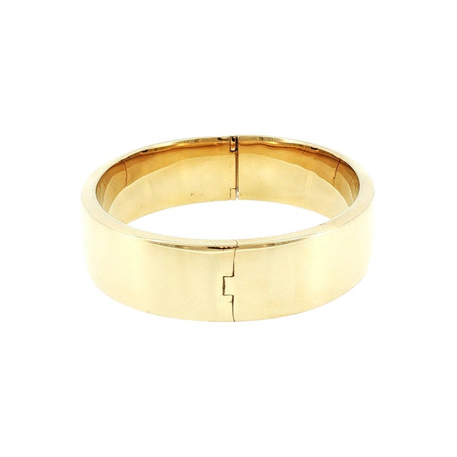 A late 20th-century 14K gold bangle bracelet. The piece is sleek and polished, measuring 15.5mm across. It is hinged with...