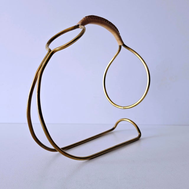 Brass wine rack in the Mid-Century Modern style with a woven raffia handle. This bottle rack is made of brass and features...