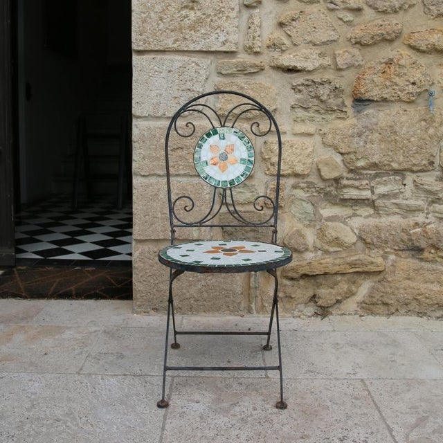 Garden antiques. folding wrought iron garden chair. It features a mosaic with a white, green, and orange floral pattern...