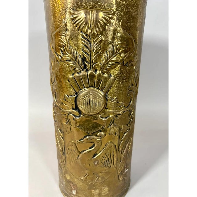 A distinctive vintage brass umbrella stand featuring richly embossed heraldic-inspired decoration, scrolling foliage, and...