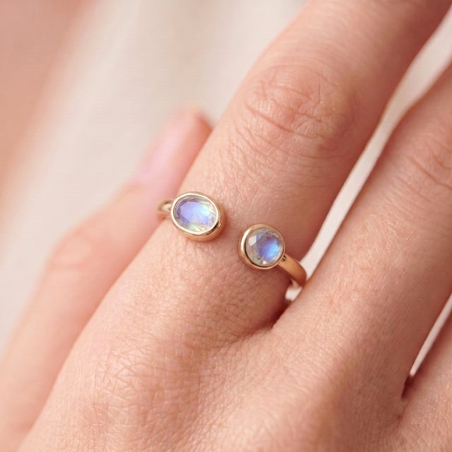 A bold, contemporary open-cuff ring featuring two luminous moonstones — an oval-cut stone paired with a round-cut stone —...